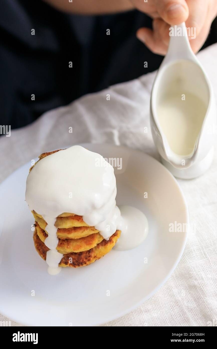 A circumcised man in a black T-shirt holds a white gravy boat in his hands and pours the curd cheesecakes on a plain white tablecloth with sour cream Stock Photo