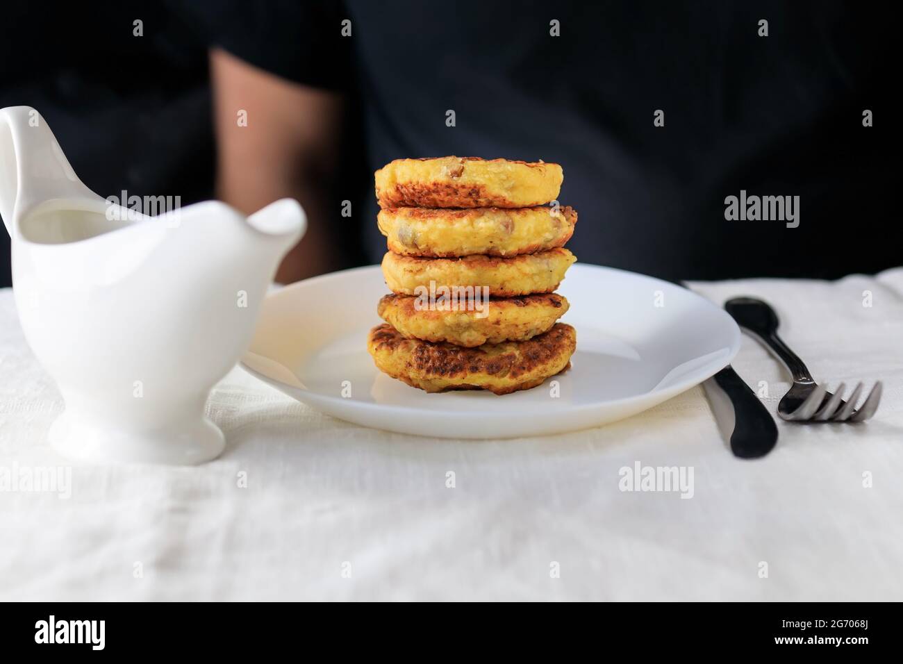A cropped man in a black T-shirt with a table on which stands a white gravy boat with sour cream sauce, curd cheese pancakes in a plate, fork, knife o Stock Photo