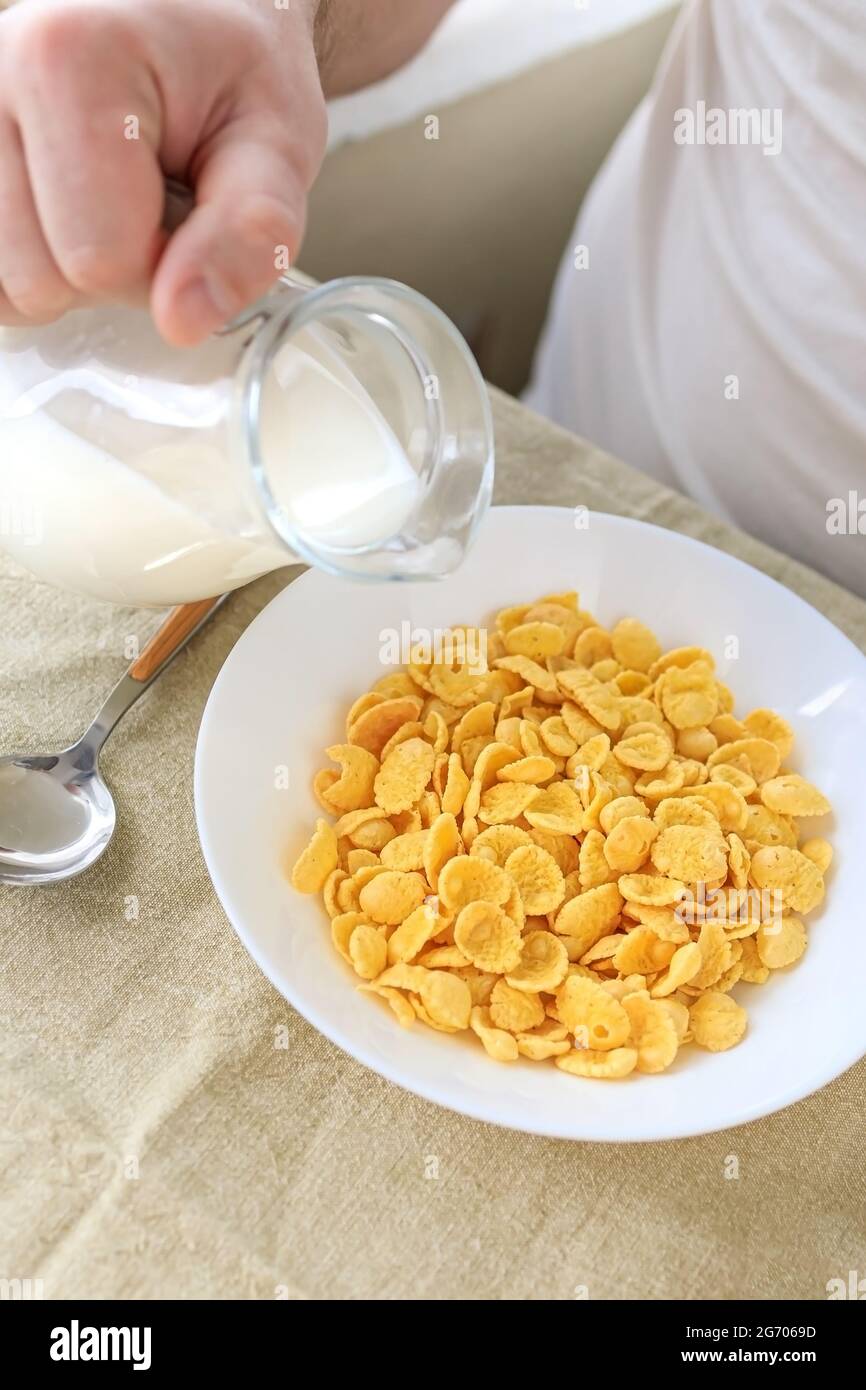 Cropped man pours milk into cornflakes in a white plate on a plain rough tablecloth. View from above. Selective focus. Concept, simple fast american h Stock Photo