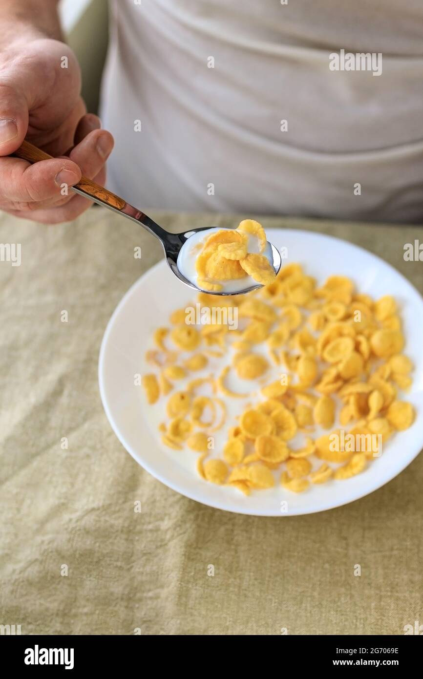 A cropped man holds a spoon in cornflakes with milk in a white plate on a plain rough tablecloth. View from above. Concept, simple fast american healt Stock Photo
