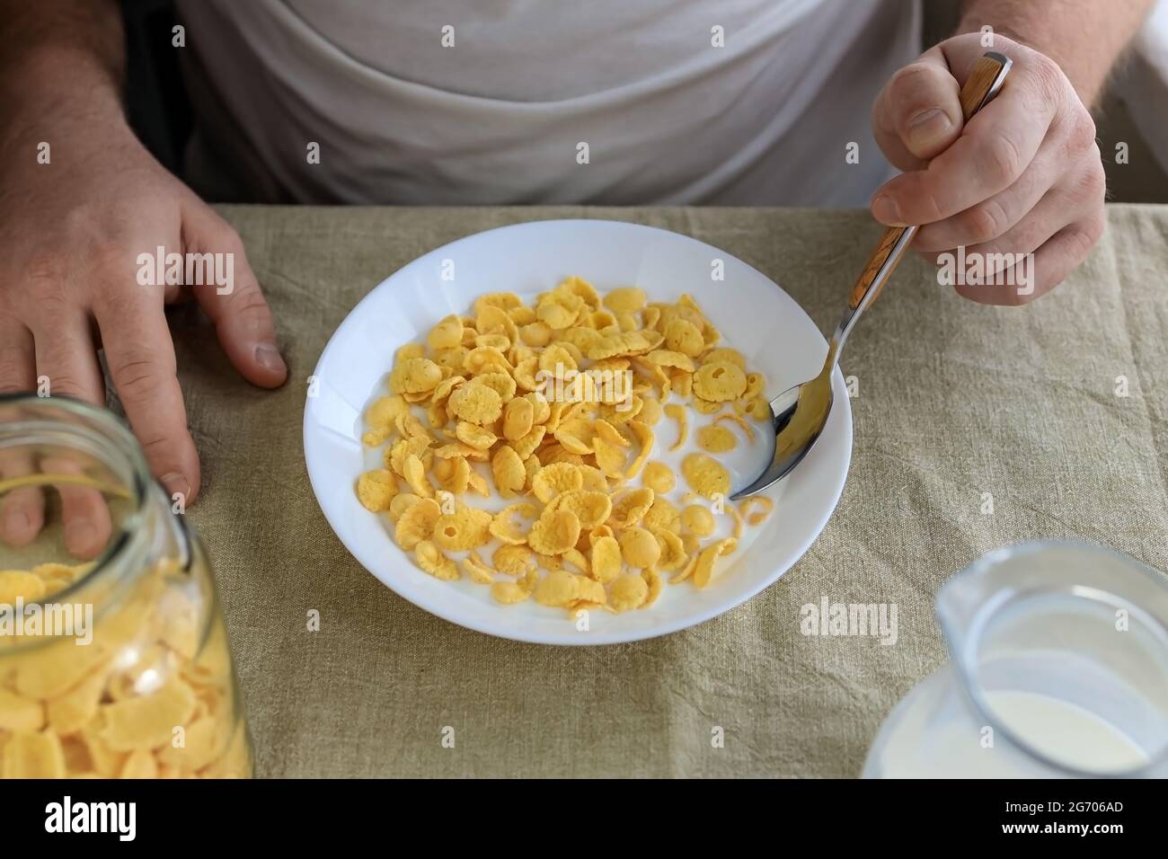 A cropped man holds a spoon in cornflakes with milk in a white plate on a plain rough tablecloth. View from above. Concept, simple fast american healt Stock Photo