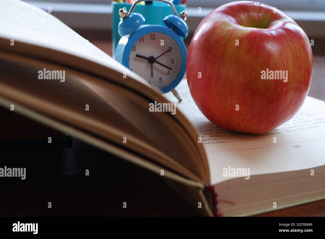 Opened book on the table near the window with an apple and an alarm clock. Stock Photo