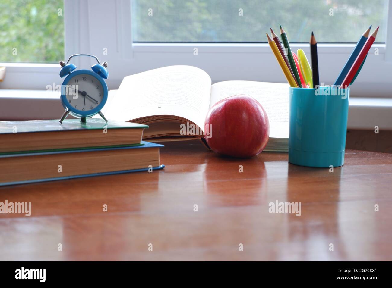 Opened book on the table near the window with an apple and an alarm clock. Stock Photo