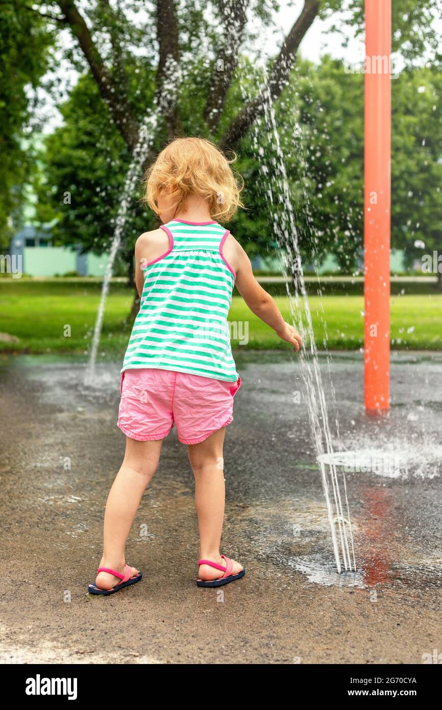 Little child playing with water at splash pad in the local public park on hot summer day. Small beautiful girl having fun at fountain playground. Stock Photo