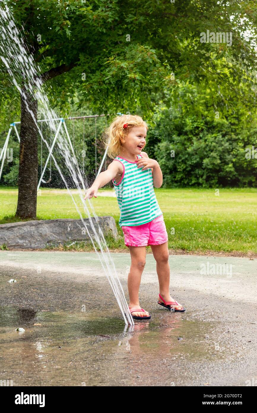 Little smiling child playing with water at splash pad in the local public park on hot summer day. Small beautiful girl having fun at fountain playgrou Stock Photo