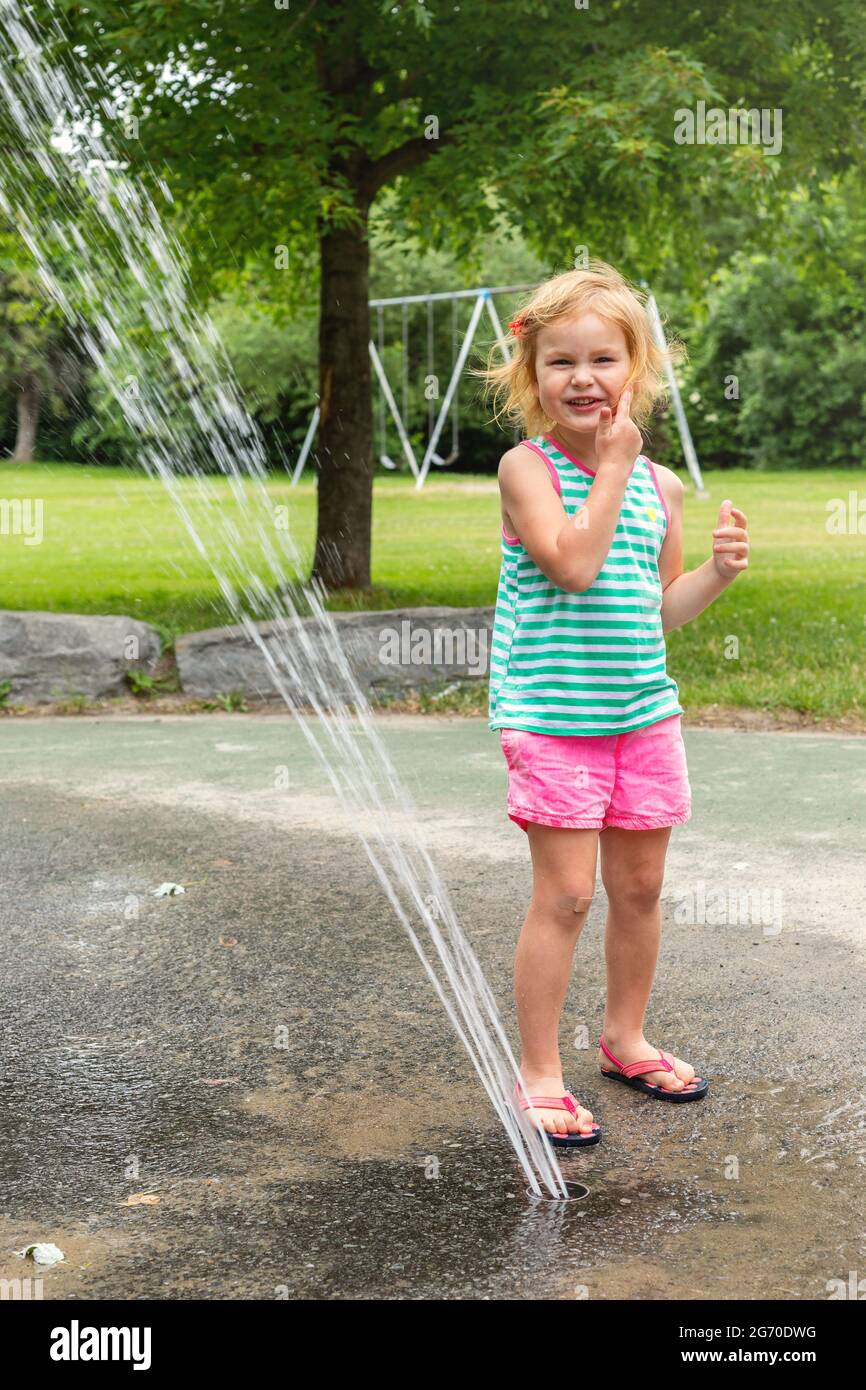 Little smiling happy child playing with water at splash pad in local public park on hot summer day. Small beautiful girl having fun at fountain playgr Stock Photo