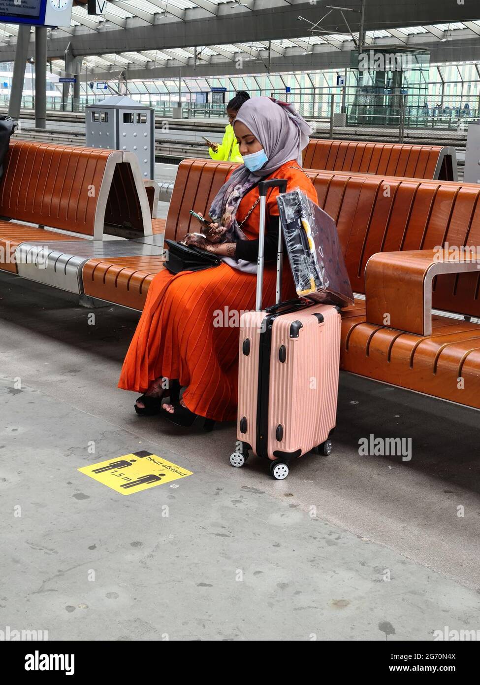 Amsterdam, Netherlands, 25th june 2021 : Lady in orange Moroccan dress wearing a scarf and mouth mask, sitting next to her luggage on a bench in the station hall of Amsterdam Bijlmer ArenA. On the ground just before her the 1,5m distance sign Stock Photo