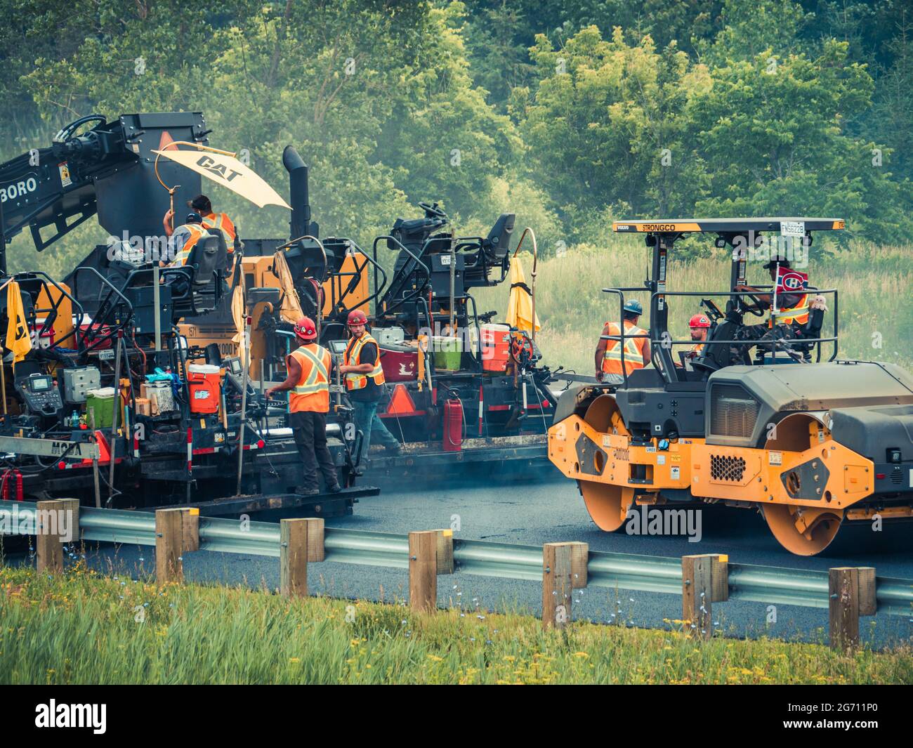 Multiple working men with hard hats and reflective orange vests operating CAT asphalt paving machinery and dump trucks on highway 25 in Quebec. Stock Photo
