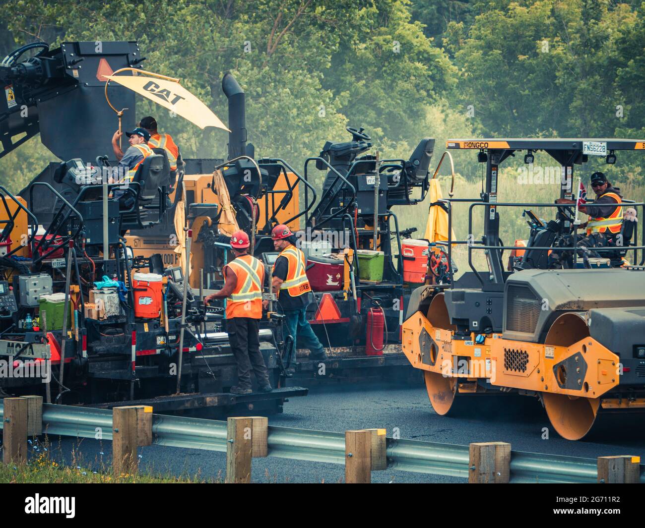 Multiple working men with hard hats and reflective orange vests operating CAT asphalt paving machinery and dump trucks on highway 25 in Quebec. Stock Photo