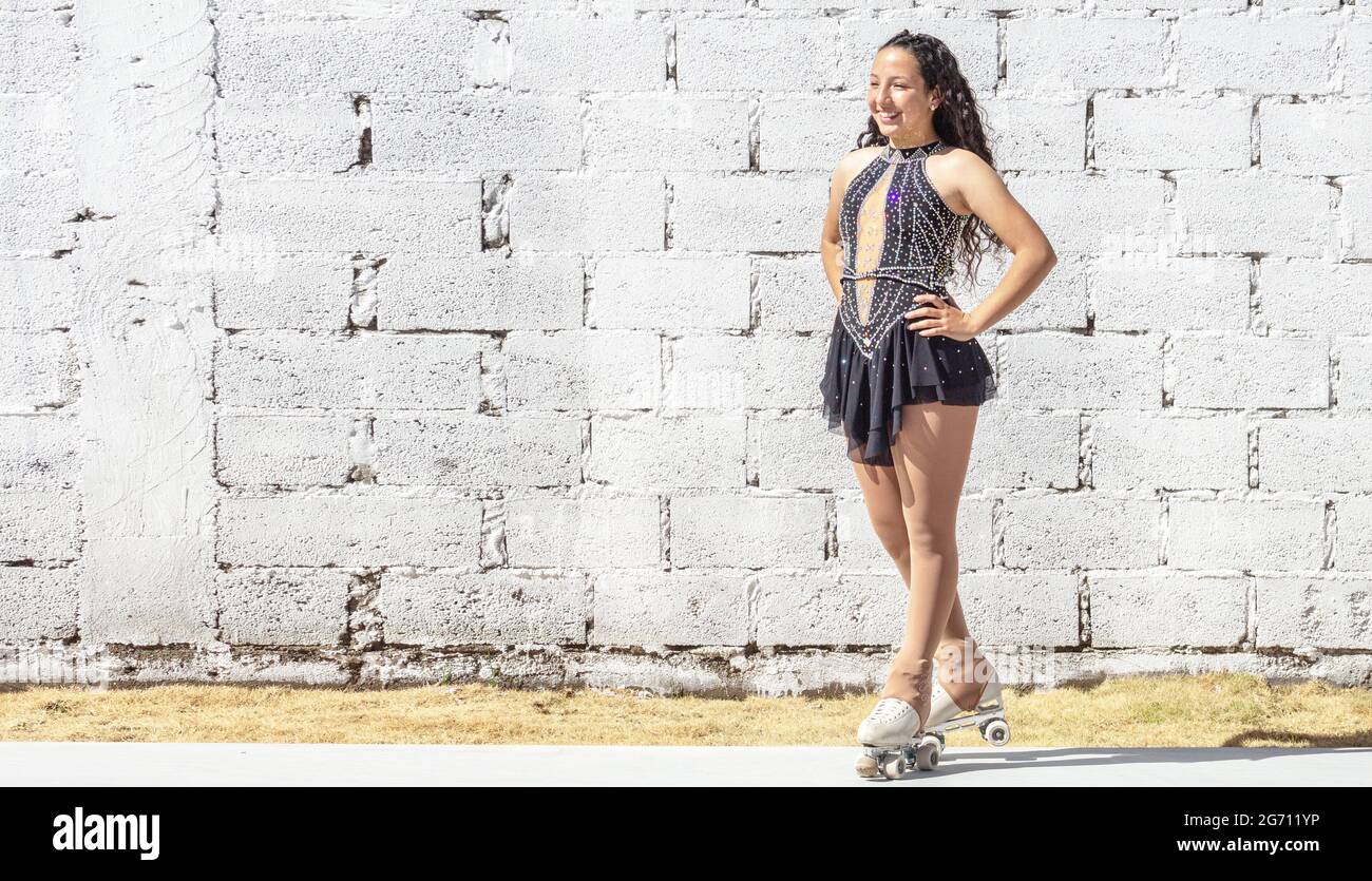 Full length portrait of Latina roller skater with curls and competition outfit outdoors smiling in front of a white block wall on a sunny morning Stock Photo