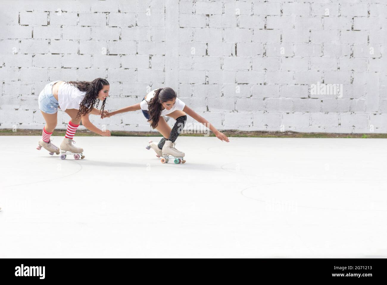 Two friends in shorts, white t-shirt and long socks skate fun on roller skates on a summer morning in front of a white block wall Stock Photo