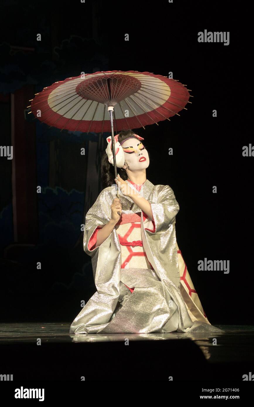 Japanese woman in traditional kimono with umbrella sitting on the knees in darkness, lighten by the moonlignt. Traditional Japanese performance. Stock Photo