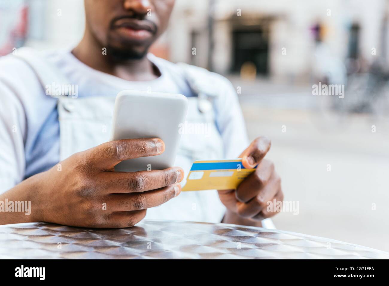 unrecognizable african young man looking to his credit card and his smart phone. He is sitting on a chair in a bar terrace Stock Photo