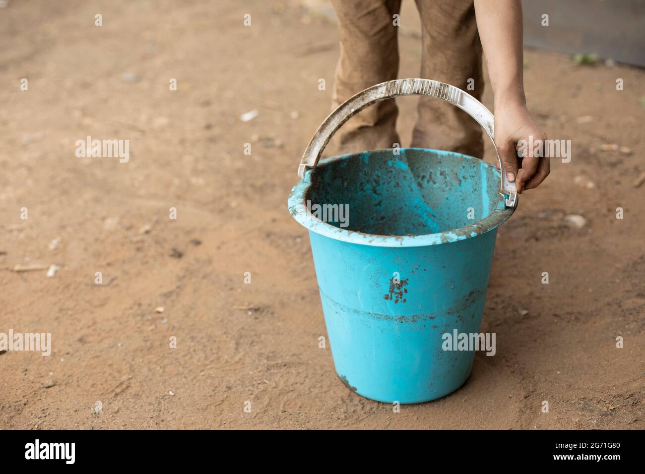 The child is holding a bucket. Blue bucket in hand. The boy plays with a bucket and a shovel. A child stained his clothes while playing in the yard. Stock Photo