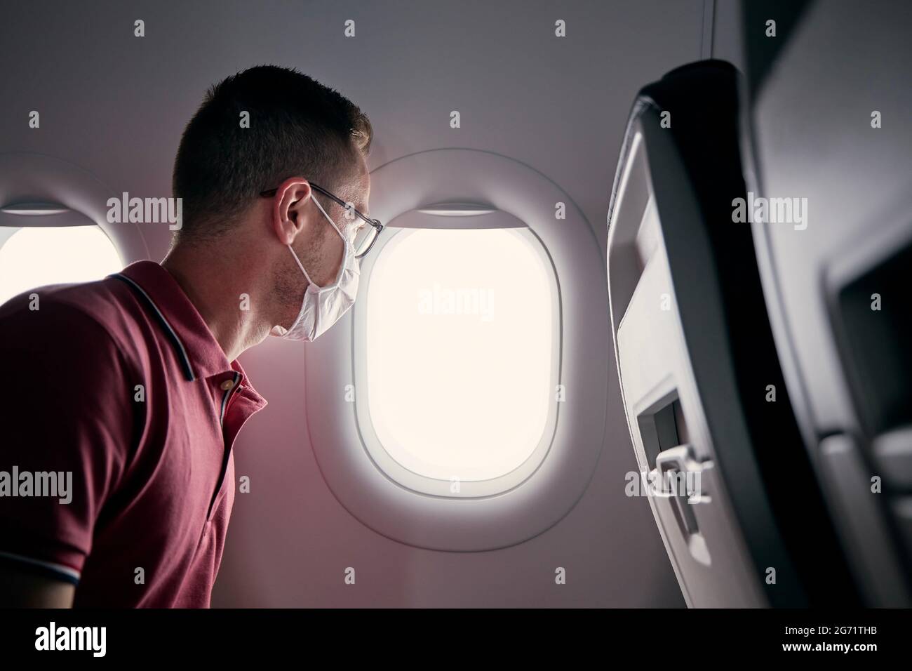 Passenger with protective face mask in airplane looking through window. Themes traveling in new normal and personal protection during pandemic covid-1 Stock Photo