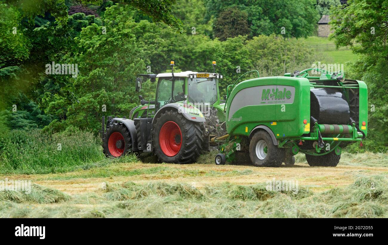 Hay or silage making (farmer in farm tractor at work in rural field, collecting dry grass, wrapped round bale in McHale baler) - Yorkshire England UK. Stock Photo