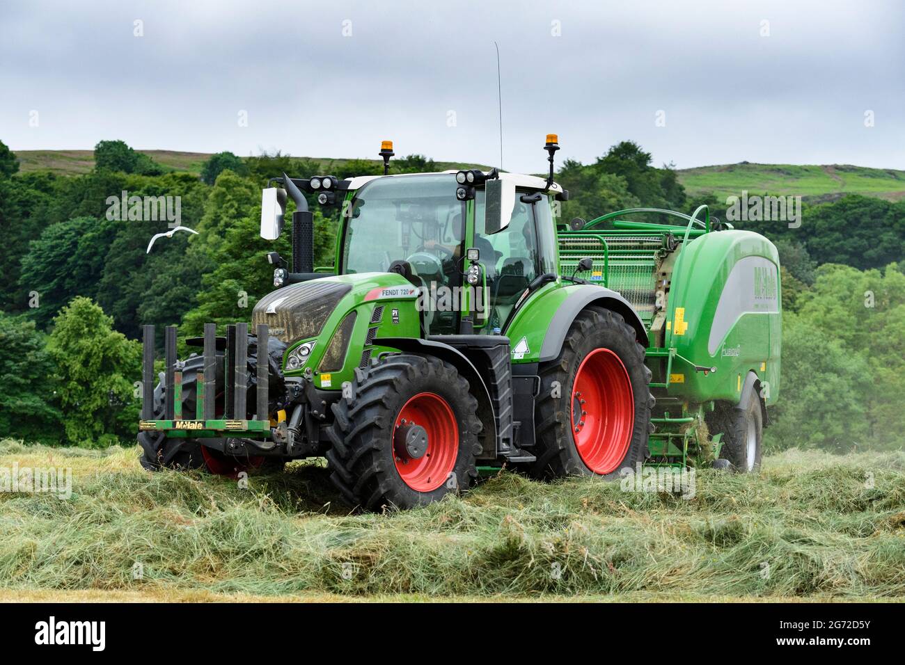 Hay & silage making (farmer in cab driving green farm tractor at work in field, pulling McHale baler, collecting cut dry grass) - Yorkshire England UK Stock Photo