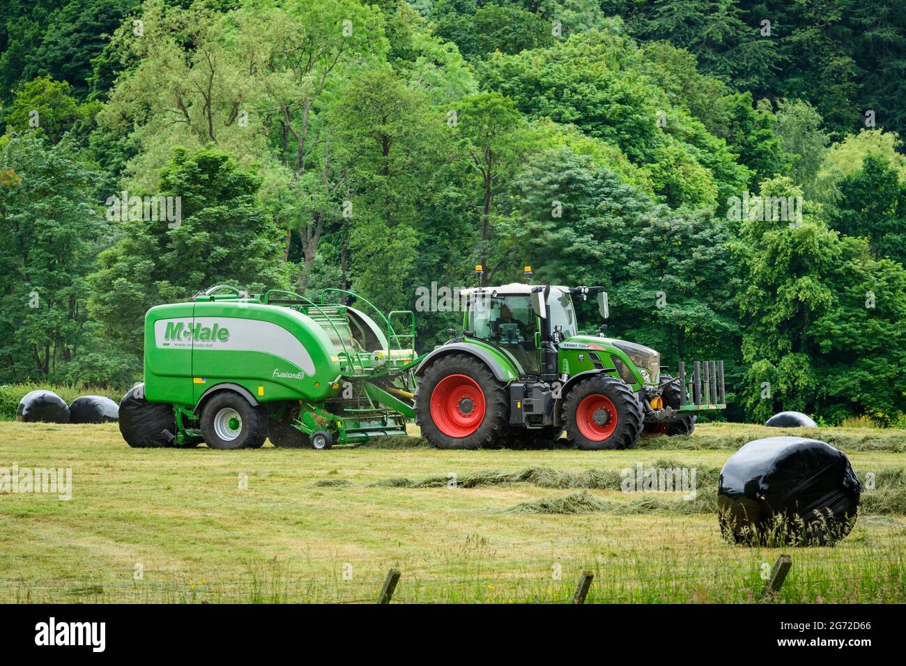 Hay or silage making (farmer in farm tractor at work in rural field pulling baler, collecting dry grass & round bales wrapped) - Yorkshire England UK. Stock Photo