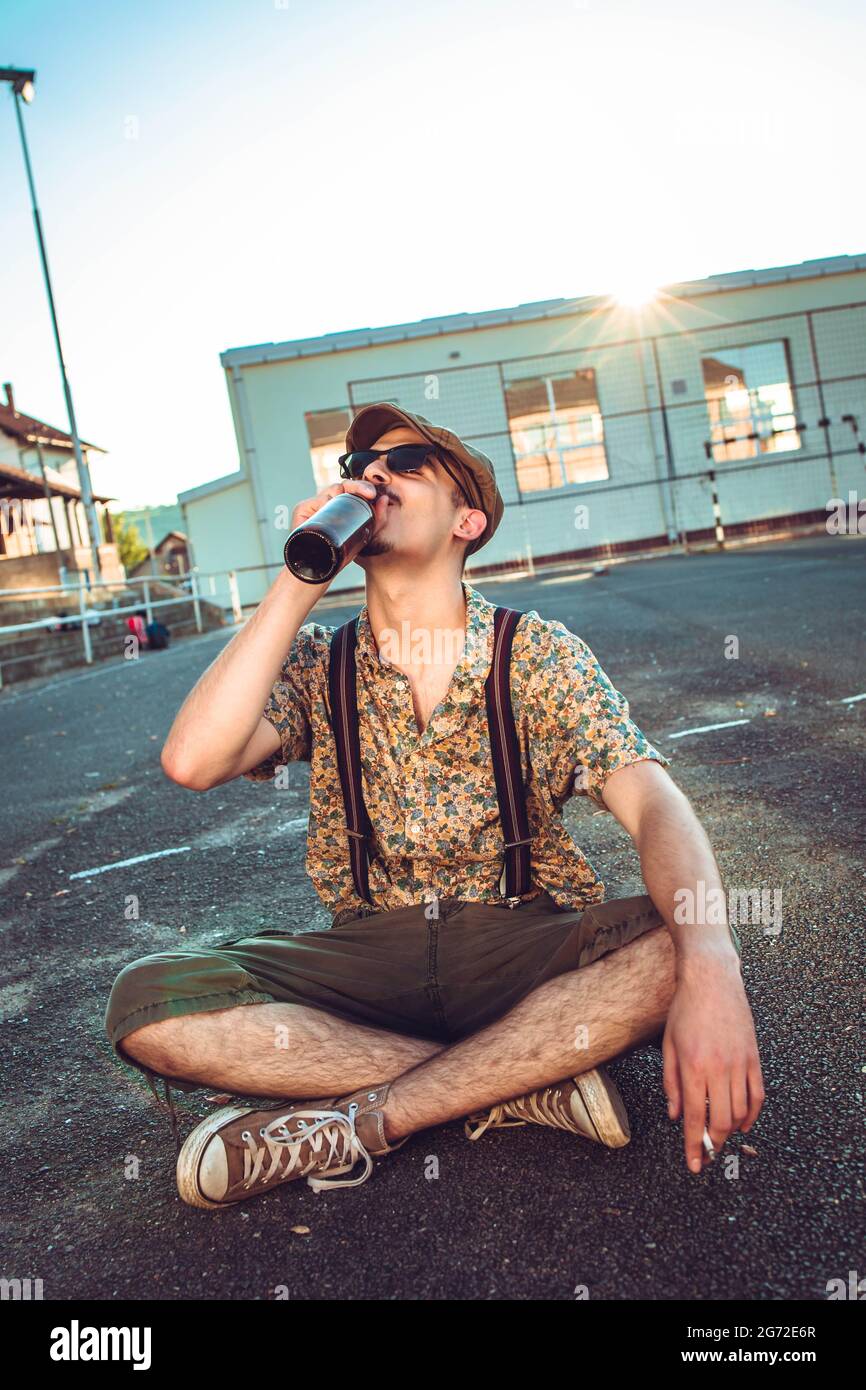 Young hipster man in retro clothing sits on the ground and drinking beer Stock Photo