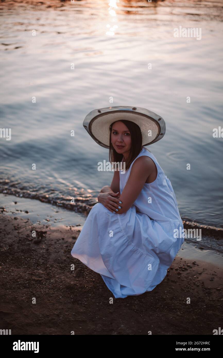 a happy Caucasian young woman in a white sundress and hat is squatting and hugging her shoulders on the beach at sunset on a summer evening Stock Photo
