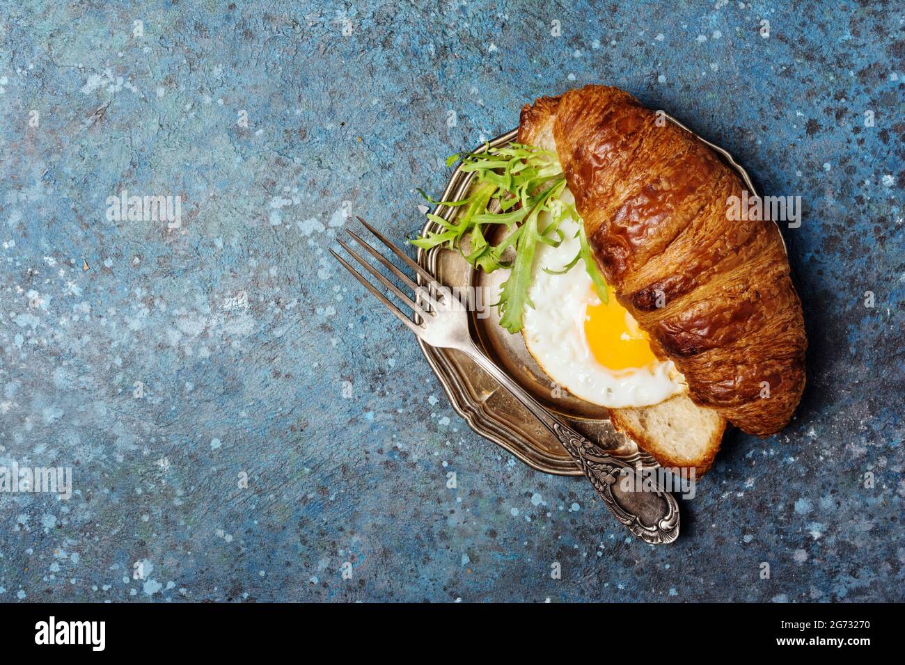 Top view of croissant sandwich with fried egg and arugula leaves on vintage silver plate and blue concrete background with copy space Stock Photo