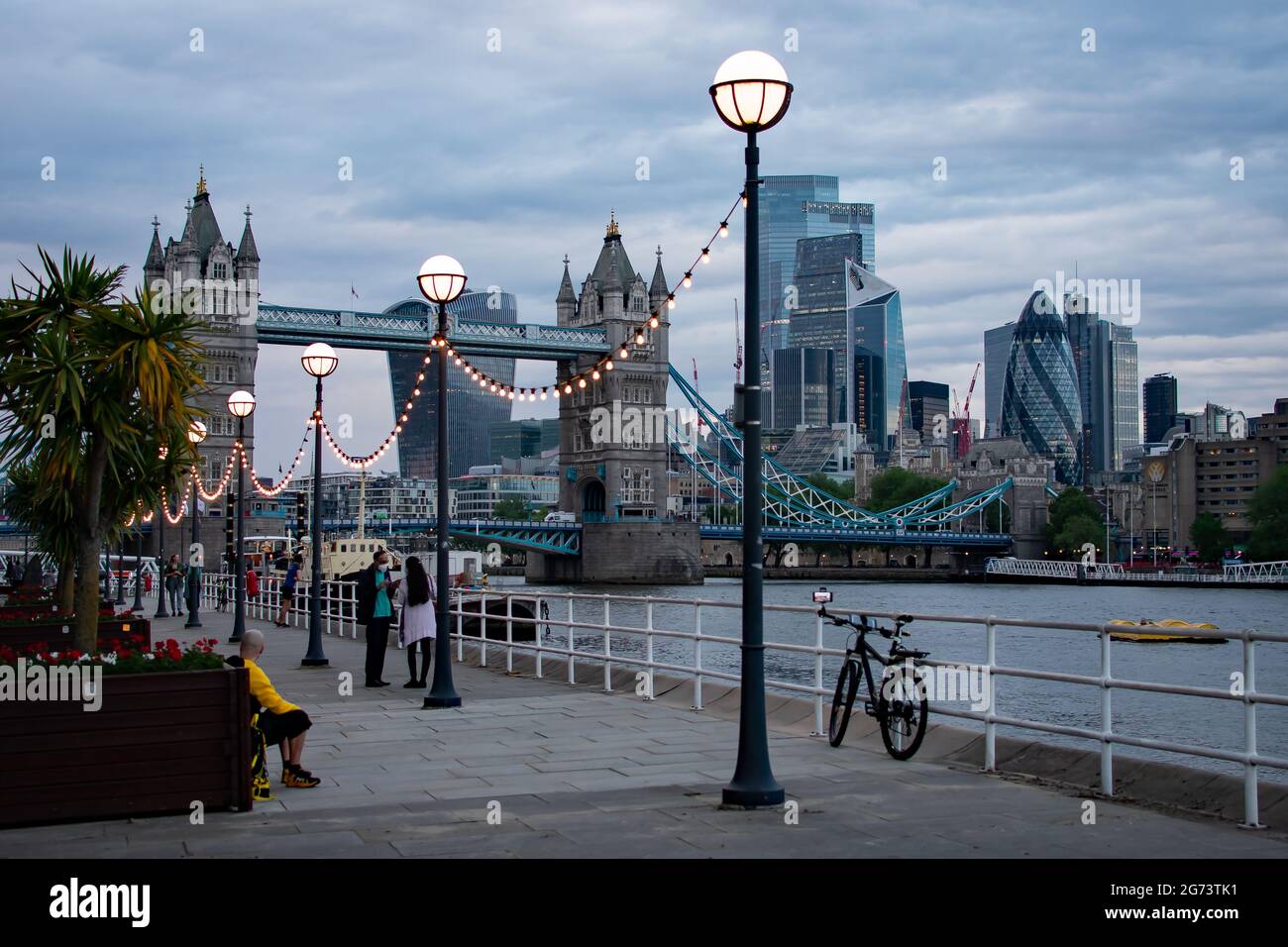 People stand along Butler's Wharf admiring the views across the City of London and Tower Bridge just before sunset as the street lamps illuminate. Stock Photo