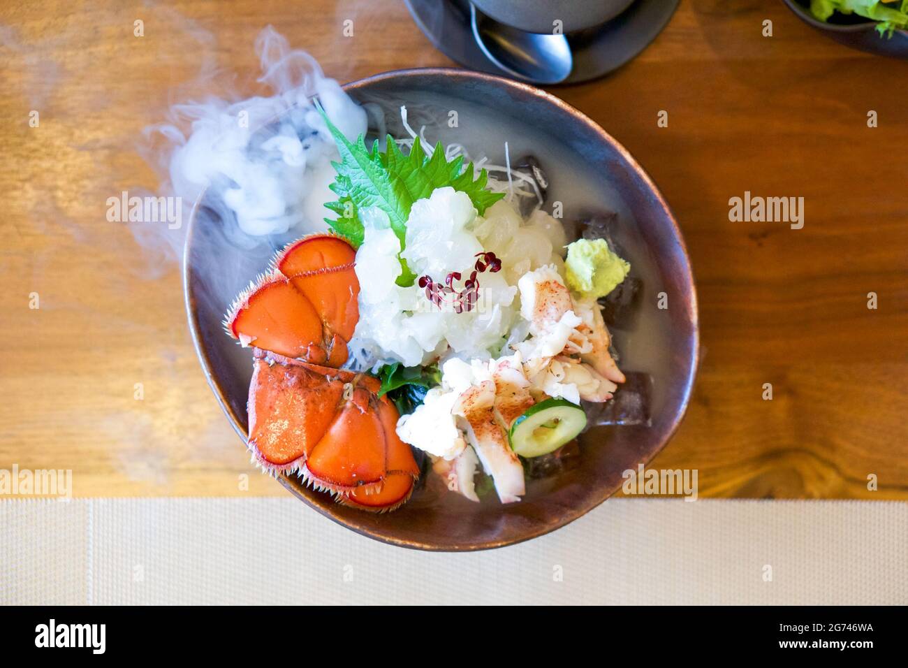 Live Canadian Lobster sashimi on brown ceramic plate with a smoke of dry ice. Served two ways fresh and boiled decorate with fresh wasabi, shredded ra Stock Photo
