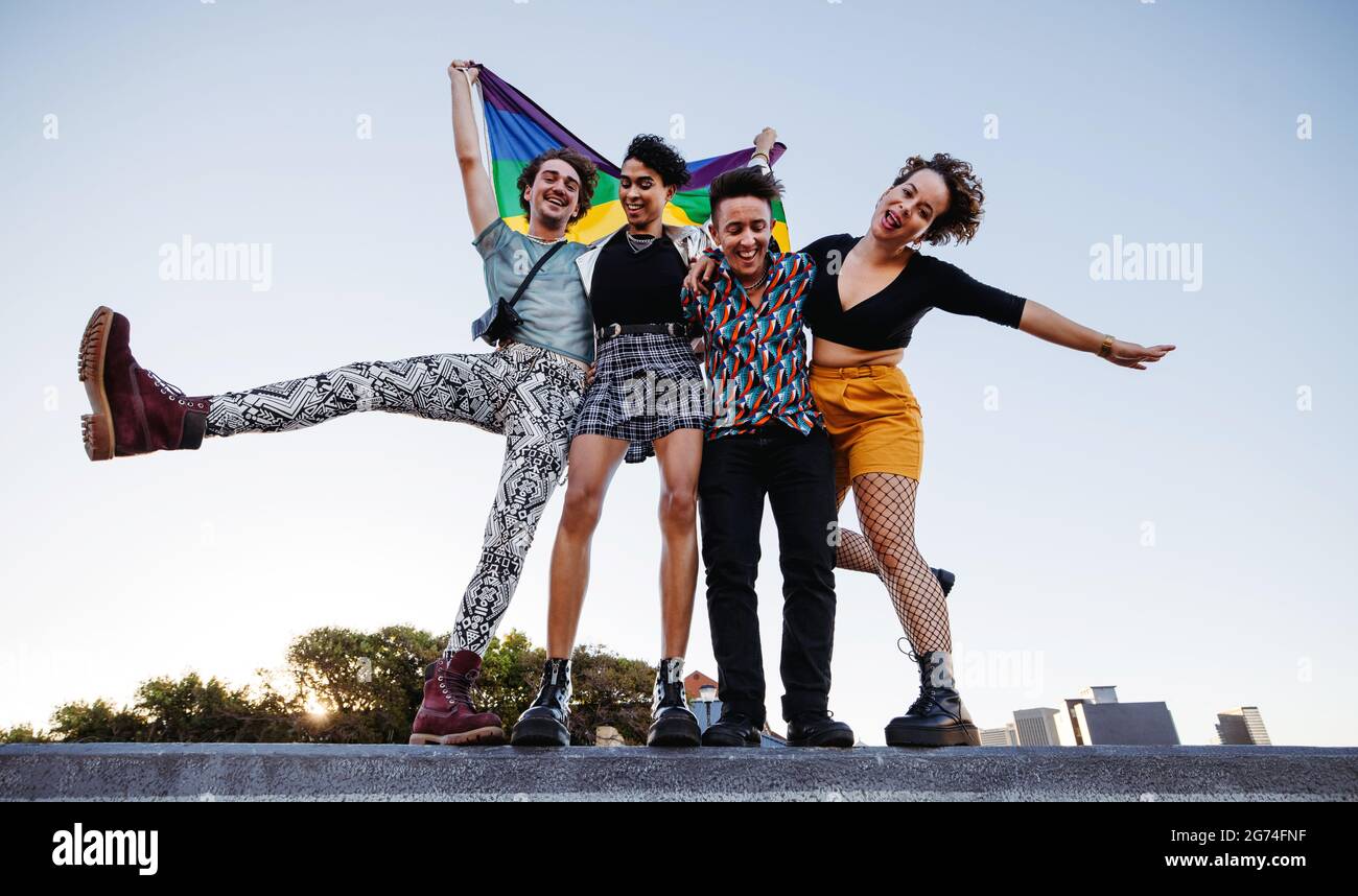Group of queer individuals celebrating gay pride together. Four members of the LGBTQ+ community smiling cheerfully while raising the rainbow pride fla Stock Photo