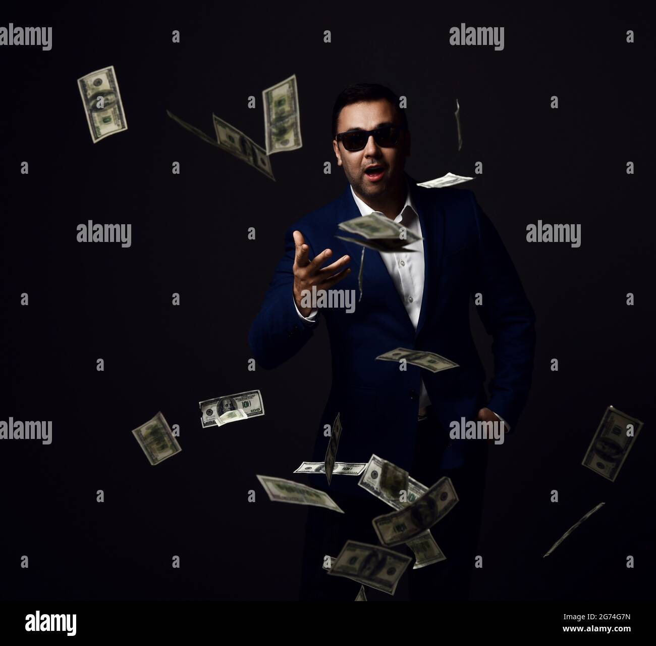 Portrait of rich successful businessman in blue jacket and sunglasses standing throwing dollars cash at camera Stock Photo