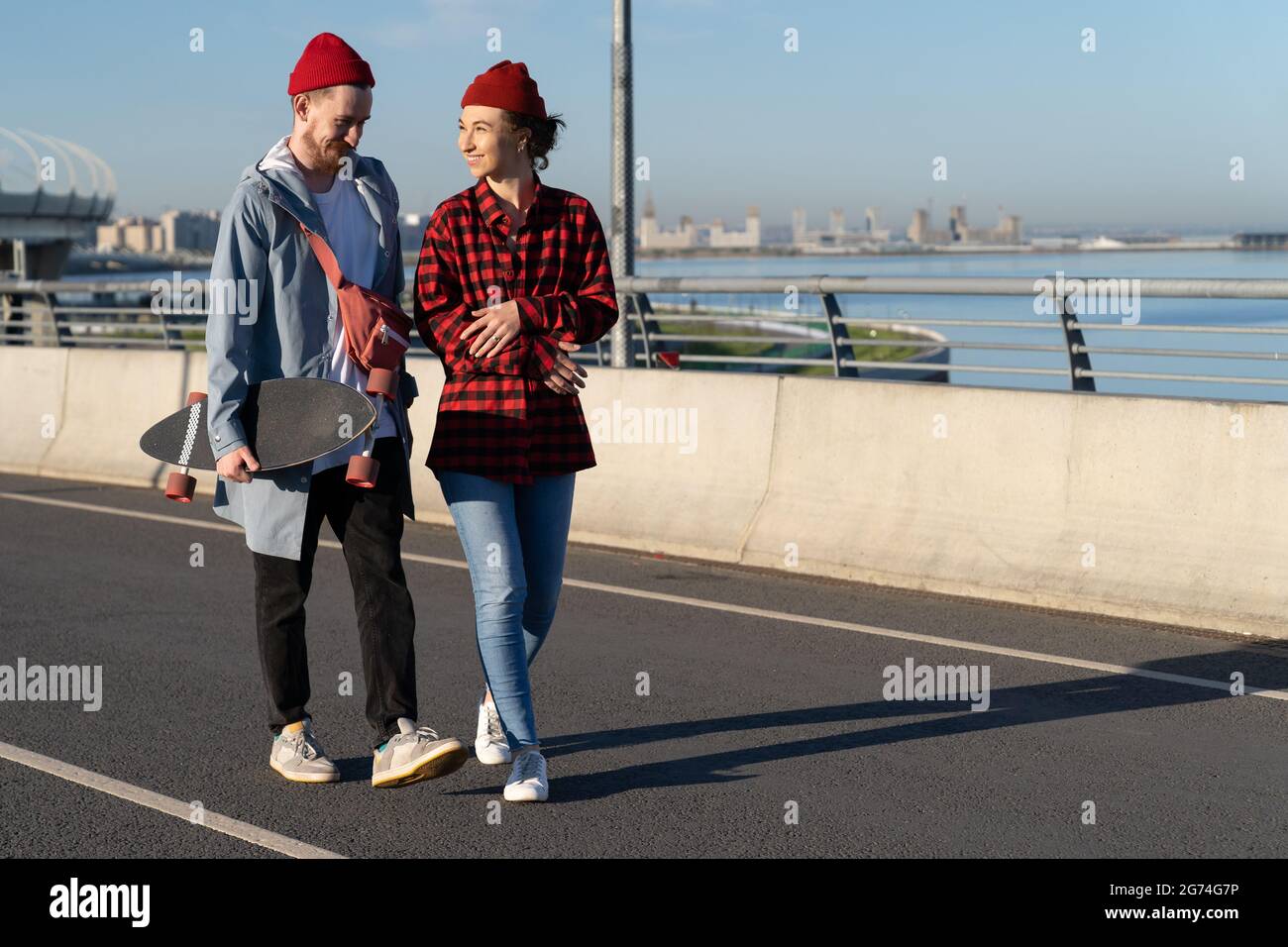 Young casual couple of hipsters walk city road carry longboard talk over urban skyline background Stock Photo