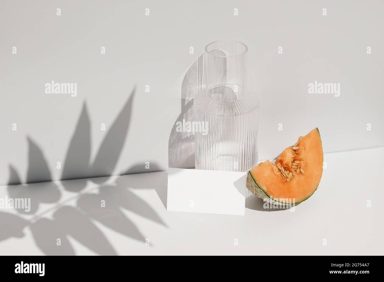 Summer food still life composition. Cantaloupe melon fruit on table. White wall with palm leaf shadow overlay. Blank paper card, invitation mockup Stock Photo