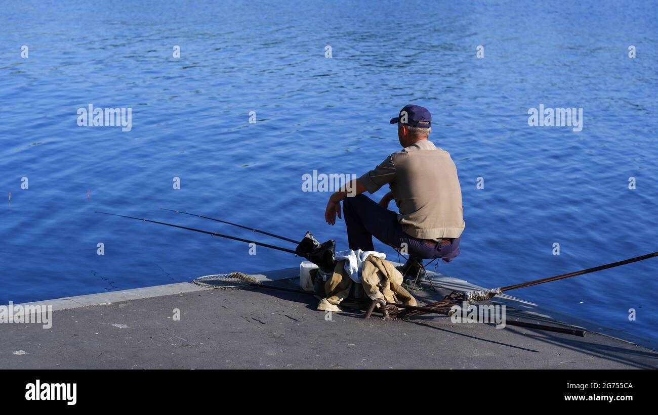 The fisherman threw two fishing rods into the water of a lake or river. The fisherman sits on a concrete pier with his back to the camera. Stock Photo
