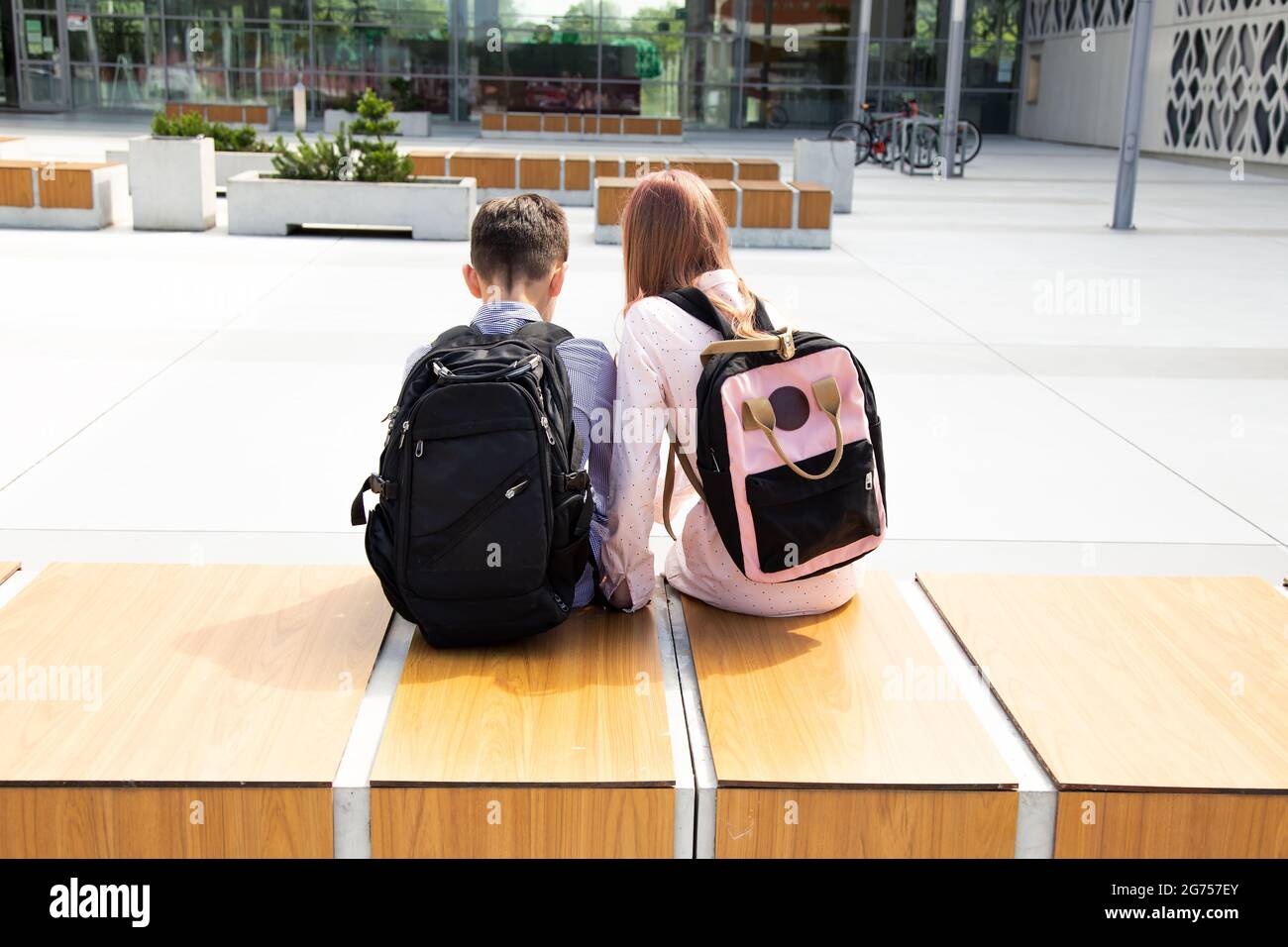 Schoolchildren girl and boy sit with school backpacks on wooden bench among concrete walls with backpacks on their backs, rear view Stock Photo