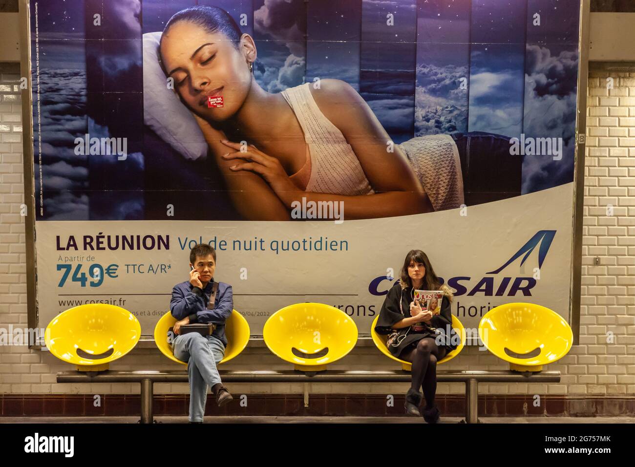 Two people seated in a Parisian metro station. Yellow seats, advertising poster in the background. Stock Photo