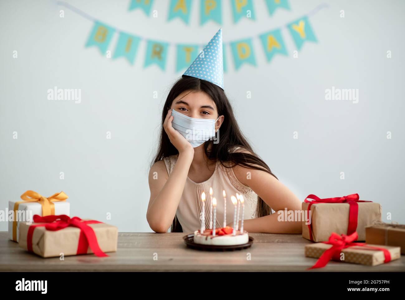 Unhappy Indian teenage girl in face mask and festive hat having dull birthday alone at home during covid isolation Stock Photo