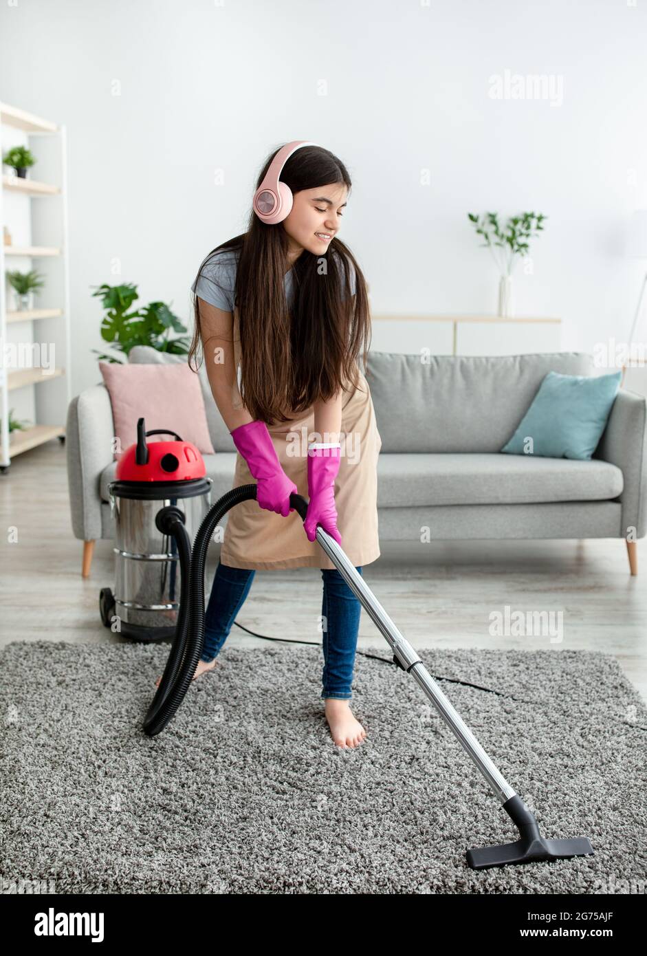 Cleaning with pleasure. Indian teenage girl vacuuming floor at home, listening to favorite music, wearing headphones Stock Photo