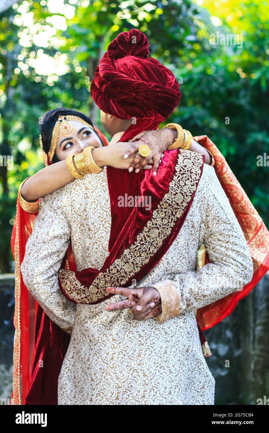 indian wedding couple hugging and groom showing victory sign with green bokeh background 2G75CB4