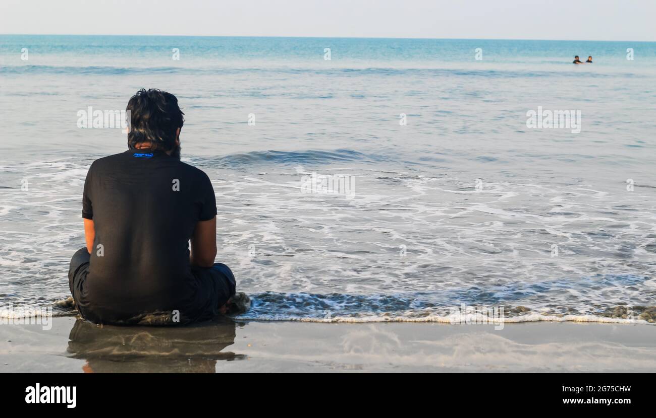 Rear view of a young man sitting on sand beach during sunrise ...