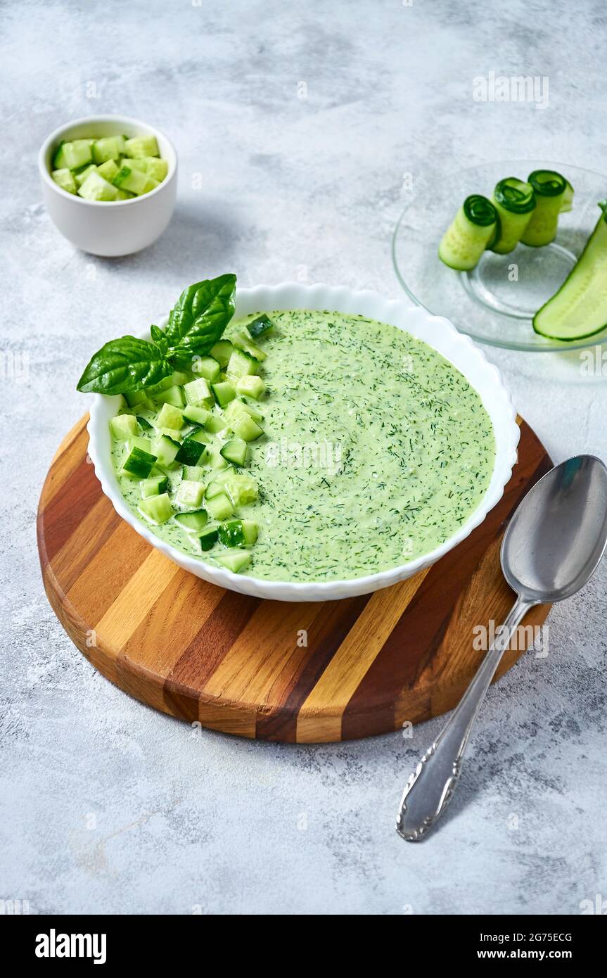 Cucumber Gazpacho - cold summer soup with basil in white bowl on wooden board on light background. Stock Photo