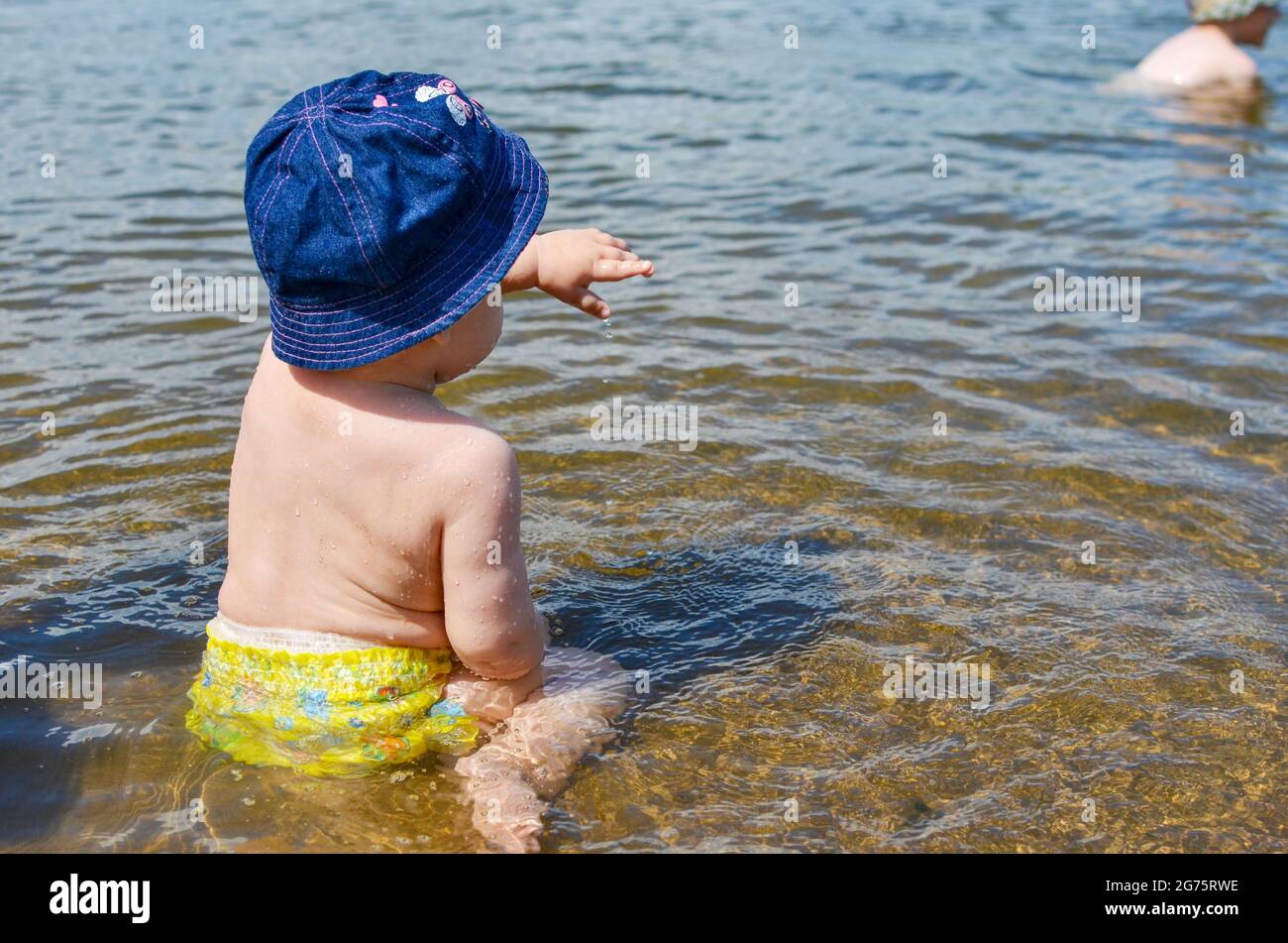little girl sits on her bottom in yellow swimming trunks and a blue panama hat in the water in the river, pointing her finger into the distance Stock Photo