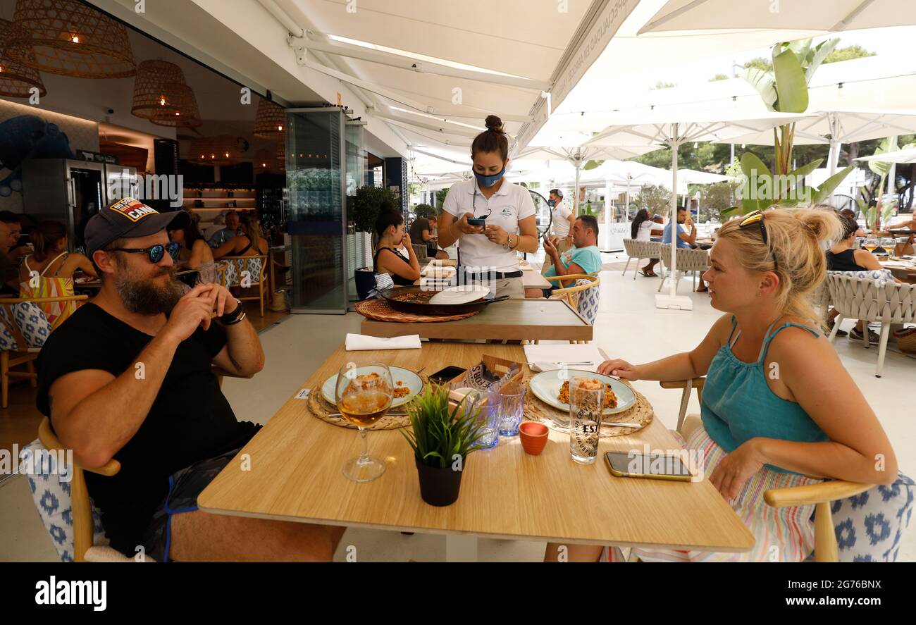 Muro, Spain. 11th July, 2021. Waitress Carmen (m) serves a paella to Polish tourists at the Can Pescador restaurant on Playa de Muro beach in the north of Mallorca. The federal government has declared the whole of Spain with Mallorca and the Canary Islands in view of rapidly increasing Corona numbers to the risk area. The practical effects for Mallorca holidaymakers are limited for the time being. Credit: Clara Margais/dpa/Alamy Live News Stock Photo