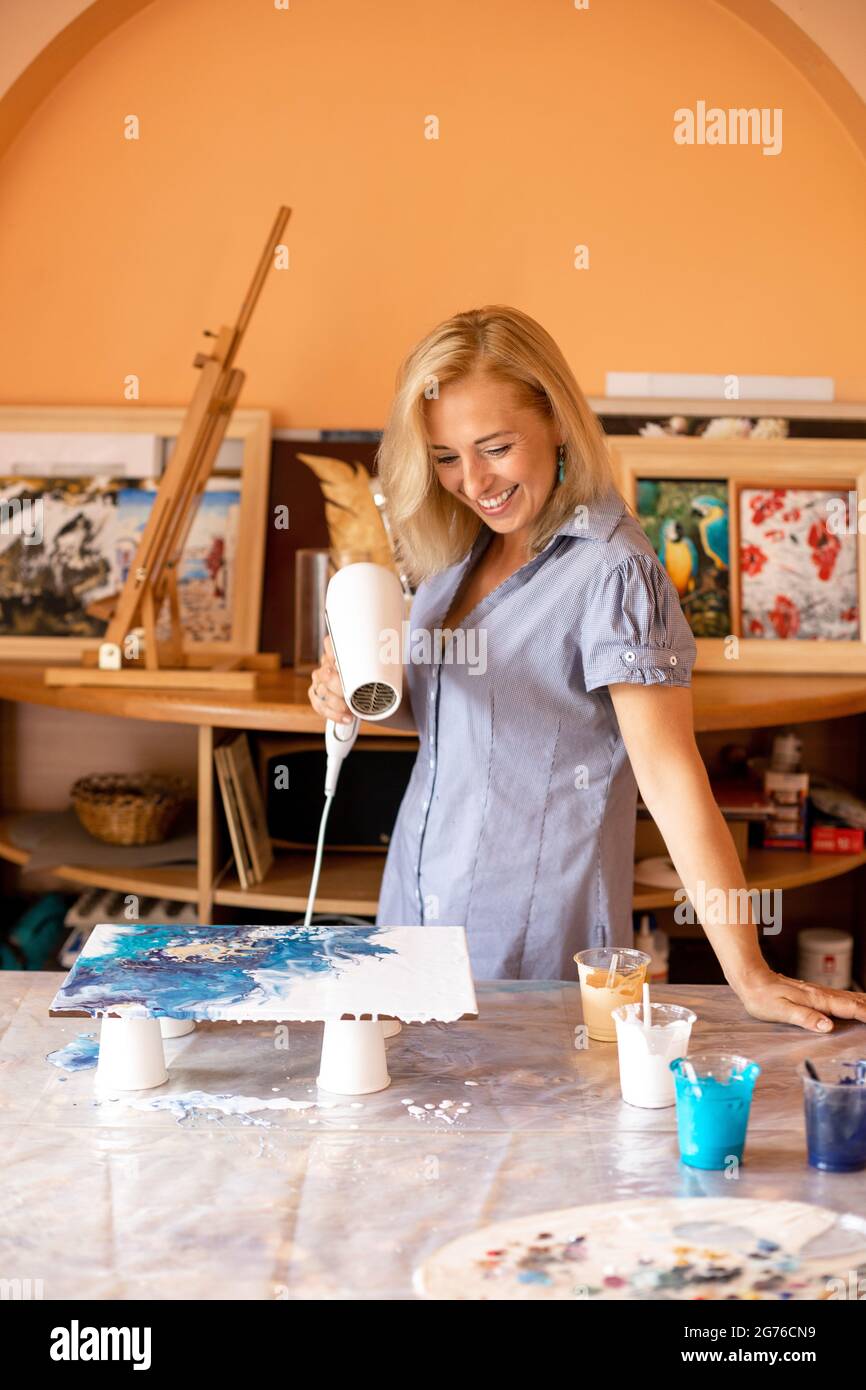 Beautiful blonde with help of paints and hair dryer creates modern interior picture, she works in her workshop. Happiness and creativity. Interior Stock Photo