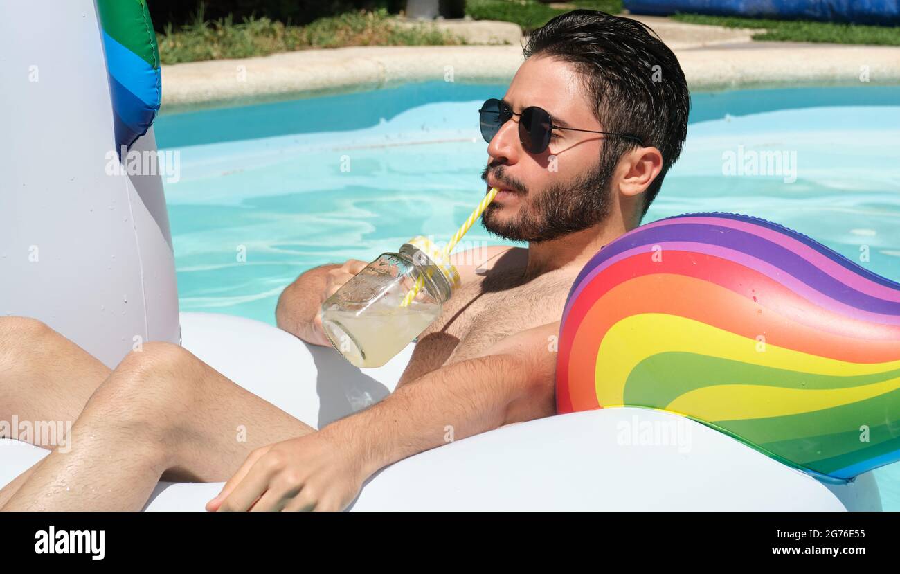 Young man wearing sunglasses drinking a refreshment on a unicorn inflatable ring in a swimming pool. Summer concept. Stock Photo