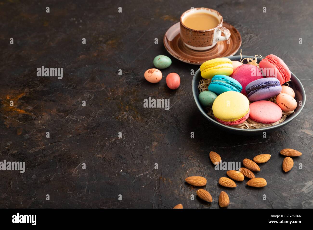 Multicolored macaroons and chocolate eggs in ceramic bowl, cup of coffee on black concrete background. side view, copy space, still life. Breakfast, m Stock Photo
