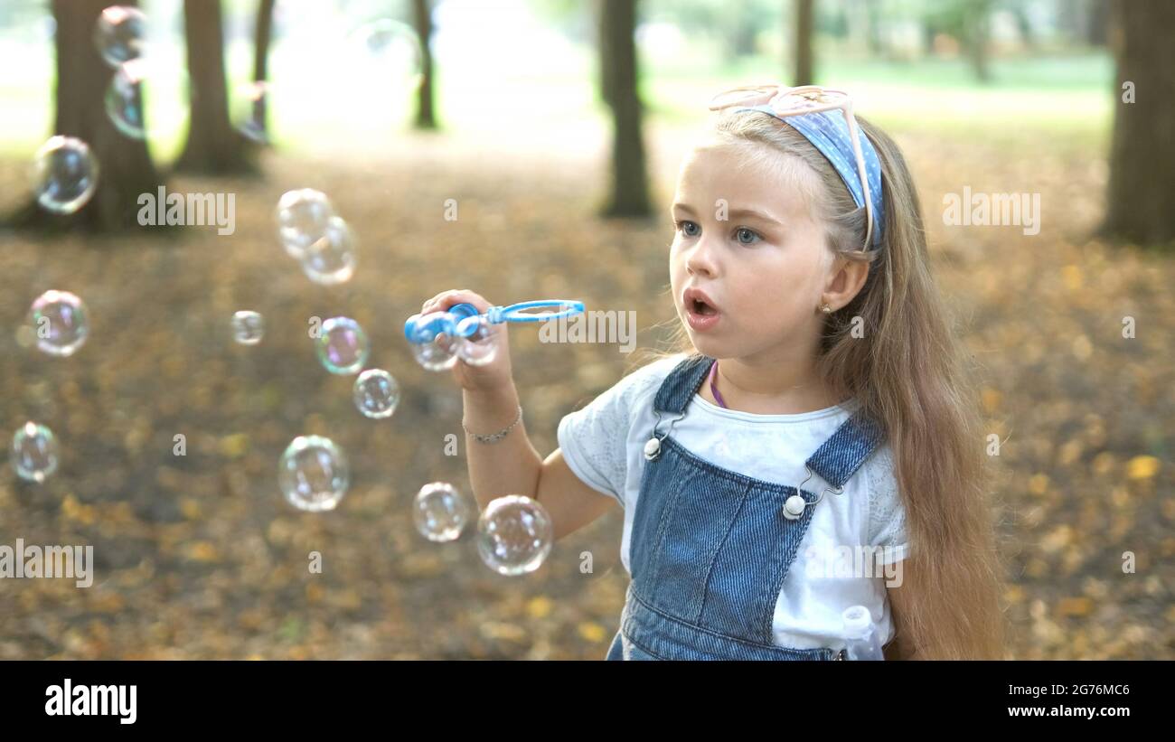 Little happy child girl blowing soap bubbles outdoors in summer park. Stock Photo