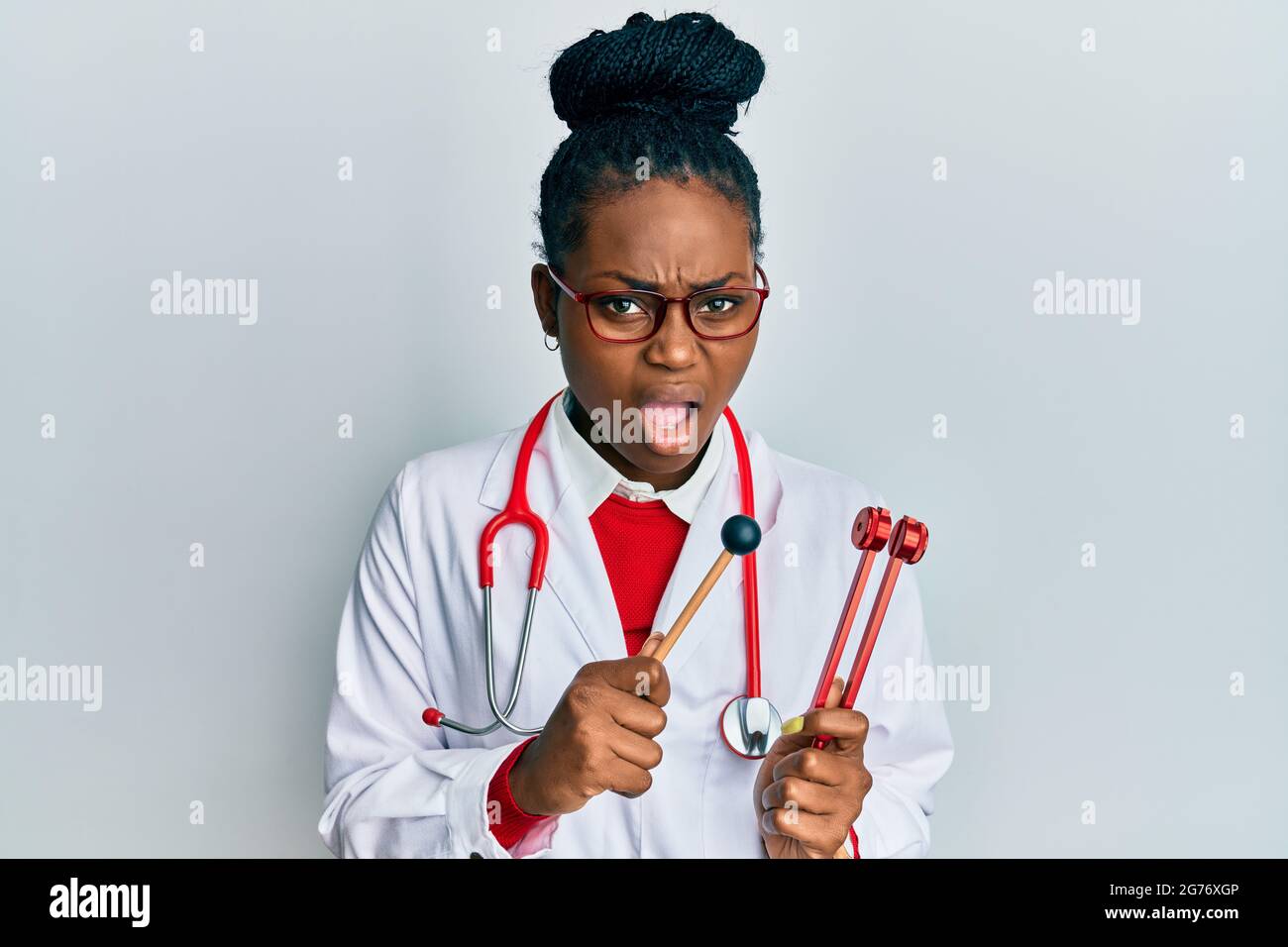 Young african american woman wearing doctor uniform holding tuning fork in shock face, looking