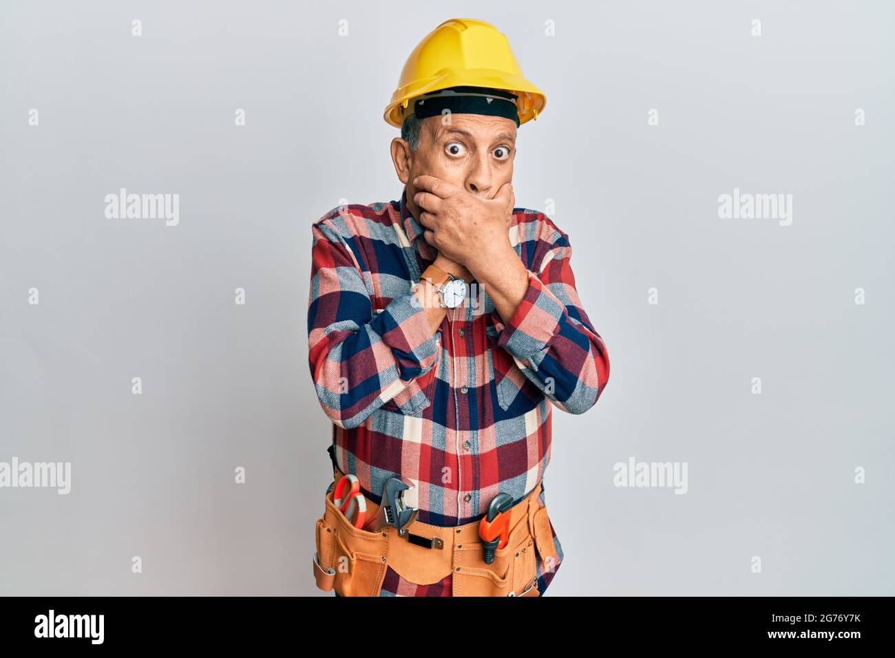 Senior hispanic man wearing handyman uniform shocked covering mouth ...