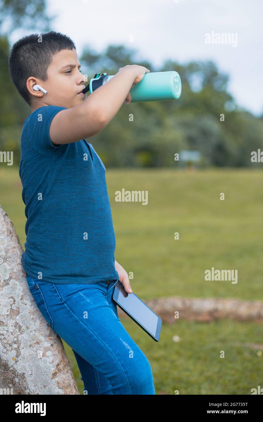 https://c8.alamy.com/comp/2G7735T/a-latino-boy-leaning-on-a-tree-in-a-park-drinking-water-from-his-bottle-with-wireless-earphones-in-his-ears-and-his-phone-in-his-hands-2G7735T.jpg