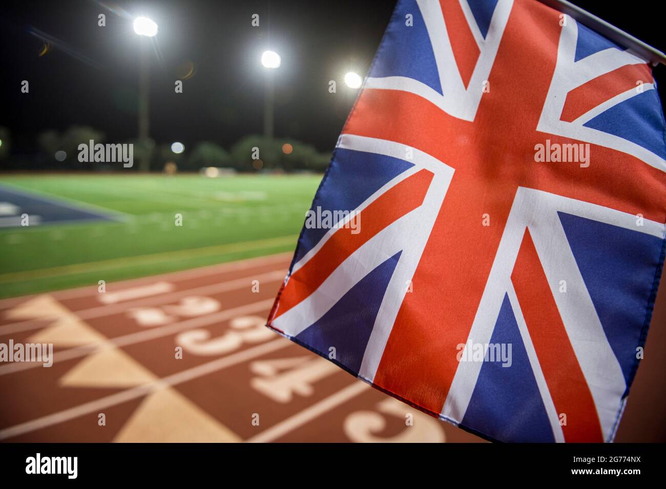 Floodlit view of UK Union Jack flying in front of the numbered lanes at the starting line of a red running track Stock Photo