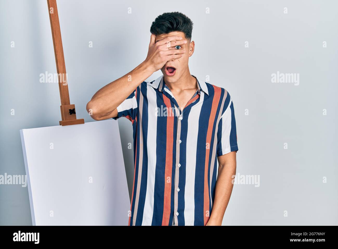Young hispanic man standing close to empty canvas peeking in shock ...